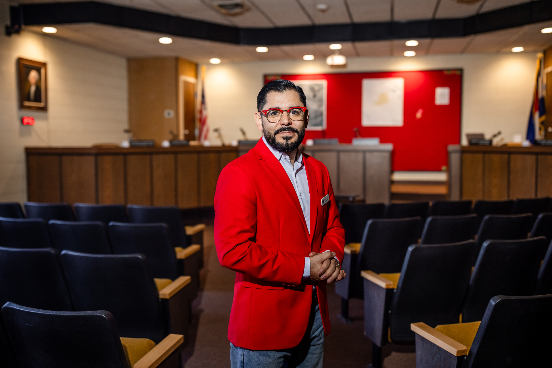 Duane Gurulé, Founder & Principal of RuralEDGE, stands in the Rocky Ford, Colorado city council chambers wearing a red blazer over a white shirt.